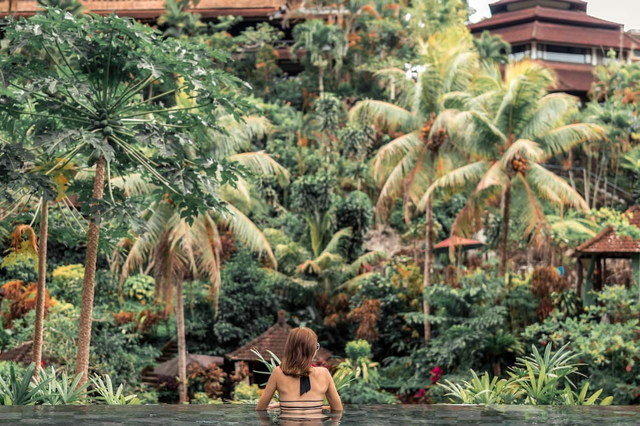 woman sitting in infinity pool overlooking forest area with palm trees and large cabins