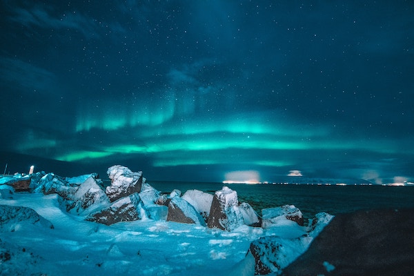 the Northern Lights glowing a blue/green color above snow covered rocks and reflecting off the cold waters