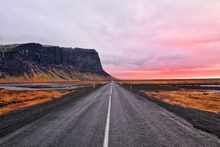 straight road leads towards steep cliff-face through orange grassland, with a orange/pink sunset in the distance