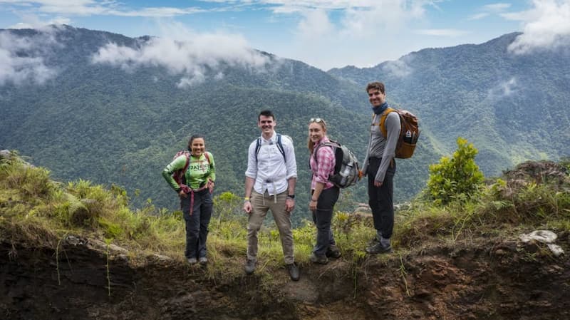 4 hikers with backpacks stood at the top of a mountain with another mountain in the distance