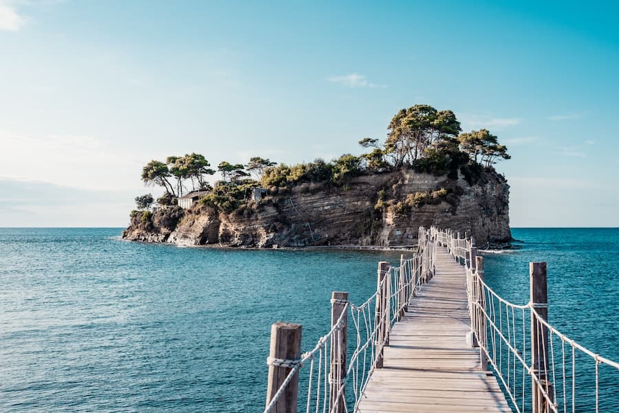 Wooden walkway with rope handrails crossing small part of the Mediterranean sea reaching tiny island with trees on