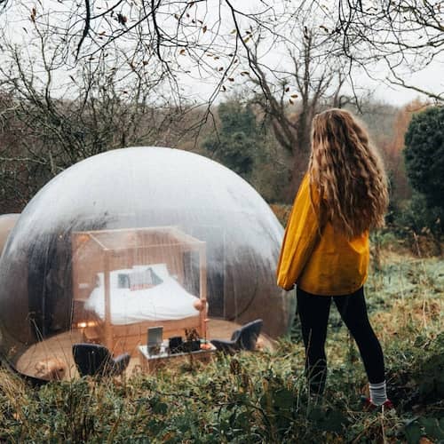 A bubble dome in the forest and a woman looking down from a hill wearing a yellow raincoat and rain boots.