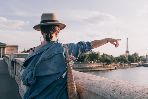 A woman wearing a sun hat looking at the Eiffel Tower from a bridge and pointing towards it.