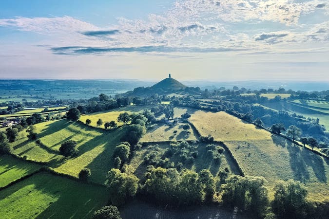 arial shot of fields in Glastonbury with sun shining and shadows cast by the trees separating the fields, tall and symmetrical hill with pillar at top stands out in middle of shot
